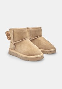 Un paio di stivaletti alla caviglia in suede beige con morbida fodera, suole in gomma piatta e un dettaglio a pom-pom testurizzato sul retro.