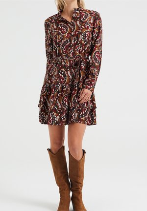 Frau trägt ein langärmeliges, knielanges Paisley-Kleid in Burgund- und Brauntönen, kombiniert mit braunen Wildleder-Kniestiefeln.