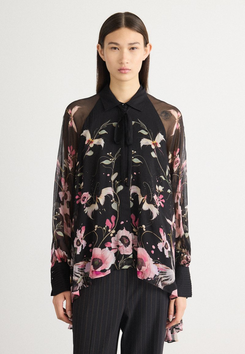 Blouse noire à fleurs avec des manches longues et transparentes ; elle présente des motifs floraux roses et blancs, un col et un nœud au niveau du cou.