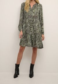 Robe verte à motif paisley avec manches longues, col tab, et jupe à volants, fabriquée en tissu léger. Portée avec des bottines noires à la cheville.