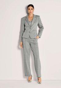 Vrouw in een op maat gemaakte grijze blazer en bijpassende broek, staande met één hand in de zak tegen een lichte achtergrond.