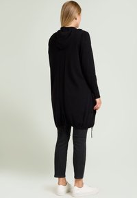 Hoodie noir long avec ourlet à cordon de serrage, poignets côtelés et coupe décontractée, associé à un jean foncé et des baskets blanches. Texture lisse.