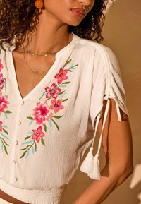 Blusa bianca ricamata con motivi floreali in rosa e verde, maniche corte con nastri e pon pon, e chiusura frontale con bottoni.