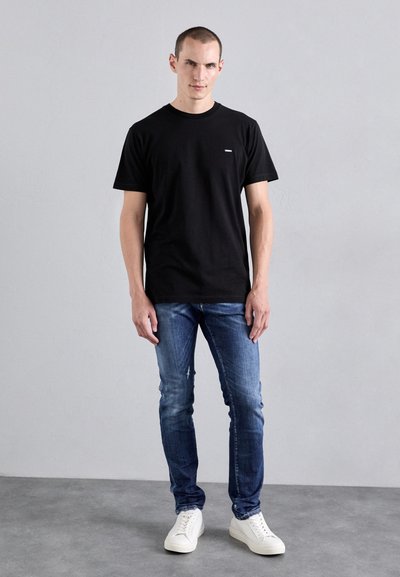 T-shirt nera in cotone con colletto rotondo, maniche corte, abbinata a jeans blu scuro e sneakers bianche. Design semplice con dettagli minimi.