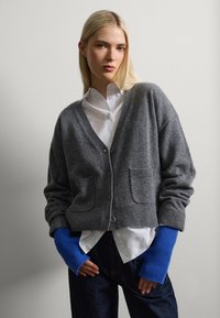 Cárdigan de lana gris con escote en V, bolsillos frontales y puños de canalé. Combinado con una camisa blanca y guantes sin dedos de color azul y de canalé.