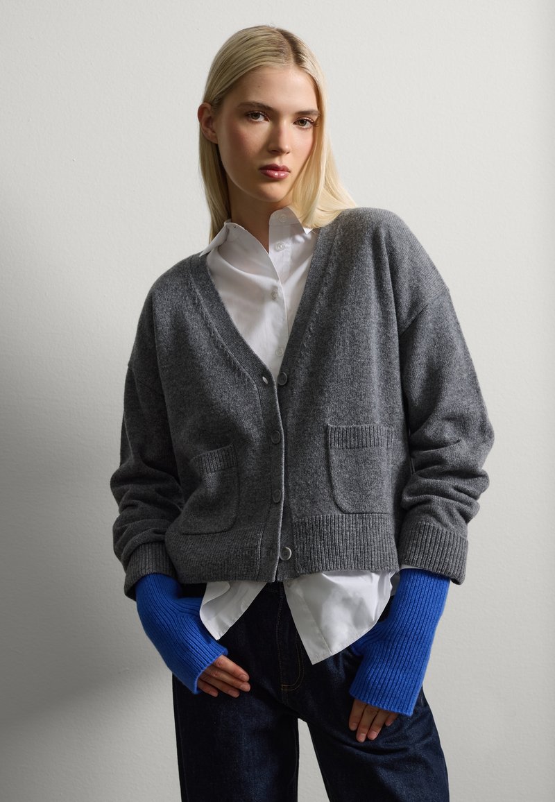 Cárdigan de lana gris con escote en V, bolsillos frontales y puños de canalé. Combinado con una camisa blanca y guantes sin dedos de color azul y de canalé.