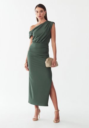 Mujer con un vestido midi verde oscuro con escote tipo off-shoulder y abertura lateral, pendientes de aro dorados y sosteniendo un bolso clutch tejido.
