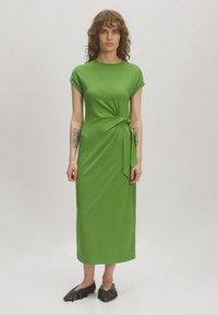 Grünes, knielanges Kleid aus glattem Stoff mit kurzen, umgeschlagenen Ärmeln und einer seitlichen Krawatte. Getragen mit schwarzen, spitzen Schuhen.