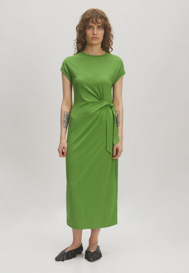 Grünes, knielanges Kleid aus glattem Stoff mit kurzen, umgeschlagenen Ärmeln und einer seitlichen Krawatte. Getragen mit schwarzen, spitzen Schuhen.