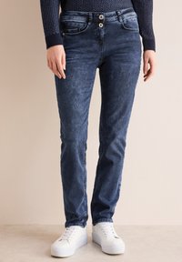 Jeans blu scuro con una trama liscia, dotati di chiusura con due bottoni, tasche laterali e lievi dettagli di sbiaditura. Abbinati a sneaker bianche.