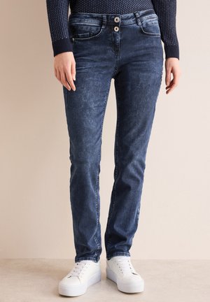 Donkerblauwe jeans met een gladde textuur, voorzien van een sluiting met twee knopen, zijzakken en lichte vervagingdetails. Gecombineerd met witte sneakers.