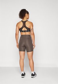 Svart sports-bh med racerback-design og utskjæringsdetalj, sammen med brune høyhøydde shorts. Modellen har på seg svarte joggesko.