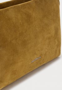 Borsa in morbido camoscio marrone con dettaglio del logo "COCCINELLE" in argento sul davanti, su uno sfondo chiaro e uniforme.