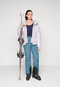 Lyslilla puffjakke, marineblå top, teal skibukser med sorte detaljer, sorte skistøvler, holder på ski. Enkel, funktionel vintersportsbeklædning.