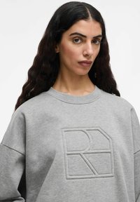 Donna con lunghi capelli scuri e mossi che indossa una felpa grigia chiara con un logo geometrico R in rilievo sul petto, sfondo bianco neutro.