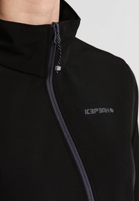 Chaqueta softshell negra con un alto cuello, cremallera frontal y acentos en gris. Presenta un sutil logo en la zona del pecho. Textura suave.