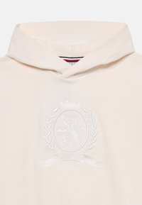 Kremfarget hettegenser med ribbestrikket halslinning, med et hvitt brodert skjoldlogo med en løve og laurbærblader i midten.