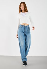 Cropped white hoodie with a zip front, paired with loose-fitting light blue jeans. The outfit is completed with black sneakers featuring white stripes.