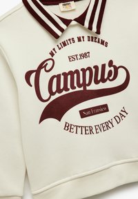 Kremowa bluza z maroonowym paskiem przy kołnierzu. Zawiera duży napis: "Campus," "EST.1987," "San Francisco," oraz "BETTER EVERY DAY." Miękka tekstura.