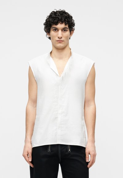 Homme aux cheveux bouclés portant un débardeur blanc à bord avant effiloché et un pantalon noir, debout devant un fond uni.