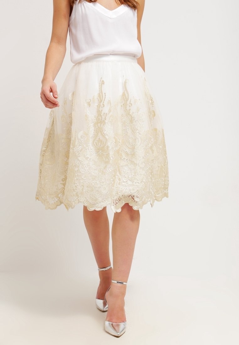 Vrouw in een witte mouwloze top en een knielange witte rok met gouden kantdetails, gecombineerd met zilveren enkelsandalen.