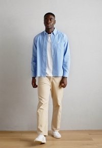 Lichtblauw overhemd met knopen over een wit T-shirt, gecombineerd met beige broek en witte sneakers. Glad textuur, casual pasvorm, minimalistisch ontwerp.
