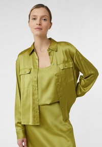 Woman wearing a shiny olive-green satin shirt over a matching tank top and skirt, standing with one hand on her hip against a plain background.