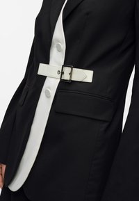 Melns žakets ar baltu apkakli, kas aprīkots ar baltu jostu ar taisnstūra sprādzi, diviem priekšējiem kabatām un pogu aizdari.