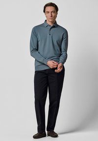 Langärmeliges Polo aus weichem blauem Stoff mit dreiknöpfiger Blende, kombiniert mit dunkler Hose und braunen Schuhen, vor hellem Hintergrund stehend.