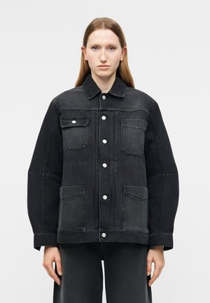 Γυναίκα με ίσια ανοιχτόκαστα καστανά μαλλιά φοράει υπερμεγέθη μαύρο τζιν μπουφάν με κουμπιά μπροστά και τέσσερις τσέπες, στέκεται μπροστά σε απλό φόντο.