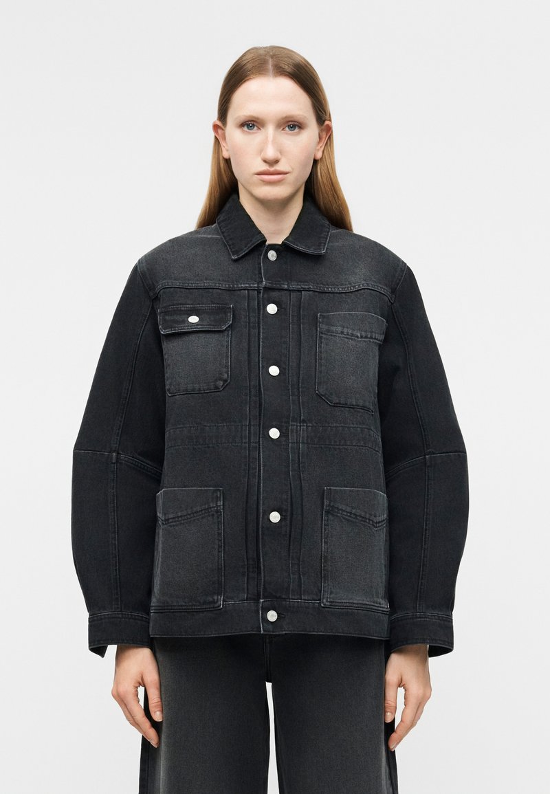 Γυναίκα με ίσια ανοιχτόκαστα καστανά μαλλιά φοράει υπερμεγέθη μαύρο τζιν μπουφάν με κουμπιά μπροστά και τέσσερις τσέπες, στέκεται μπροστά σε απλό φόντο.