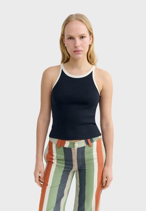 Femme portant un débardeur noir sans manches avec une bordure blanche et un pantalon à rayures verticales multicolores, debout devant un fond uni.