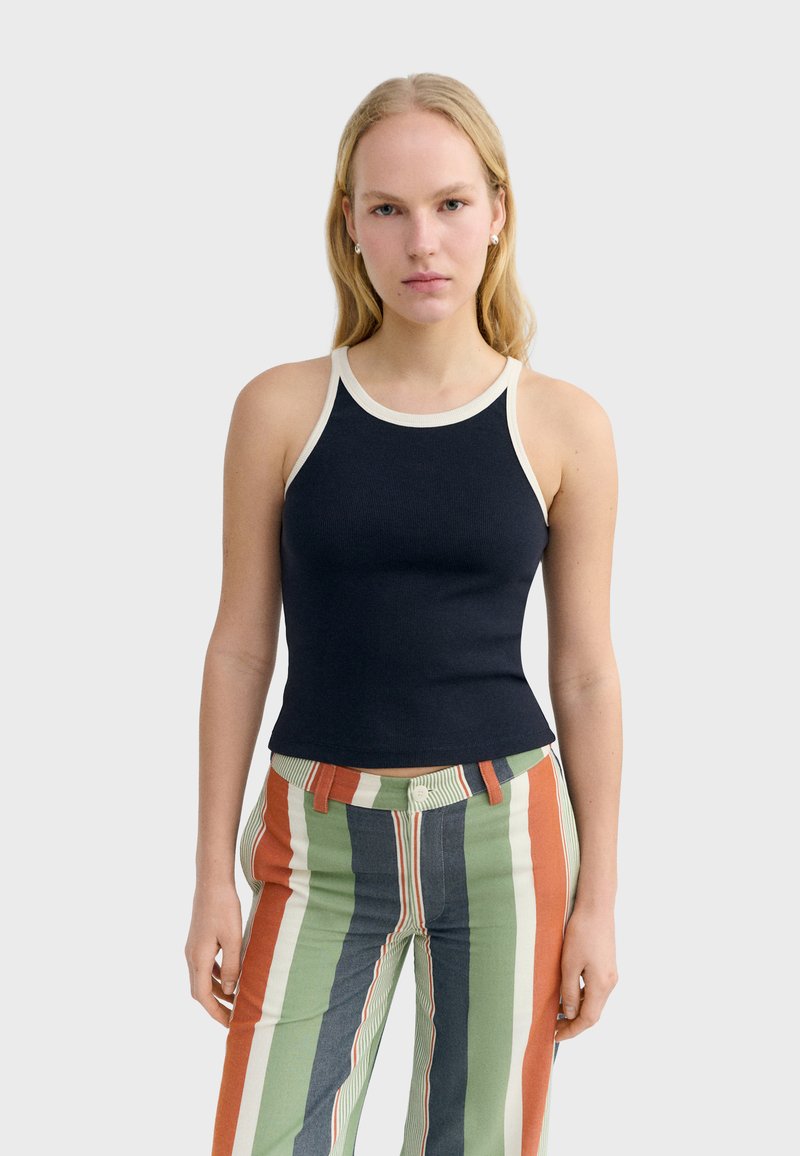 Femme portant un débardeur noir sans manches avec une bordure blanche et un pantalon à rayures verticales multicolores, debout devant un fond uni.
