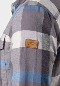 Manche d'une chemise à carreaux gris, blanc et bleu avec un patch rectangulaire marron portant l'inscription "Swedemount" avec des contours de montagnes.