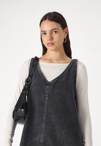 Črna denim brezrokavna obleka z V-izrezom, ki vsebuje podrobnosti šivanja in obrabljeno teksturo. Kombinirana z oprijetim dolgim rokavom in črno torbico z naramnico.