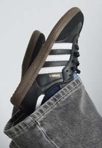 Sneaker en cuir noir avec des rayures blanches et le logo doré "SAMBIA". Comprend une semelle en caoutchouc marron, des accents texturés et un fond en denim.