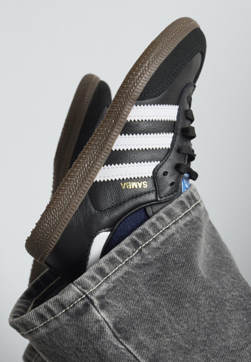 Sneaker en cuir noir avec des rayures blanches et le logo doré "SAMBIA". Comprend une semelle en caoutchouc marron, des accents texturés et un fond en denim.
