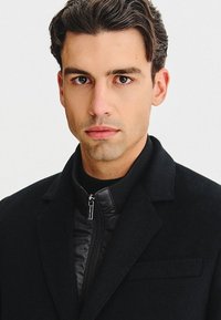 Czarny płaszcz o teksturowanej powierzchni, z mocnym kołnierzem oraz zapinaną na zamek, ocieplaną warstwą wewnętrzną. Model ma starannie ułożone włosy i neutralny wyraz twarzy.