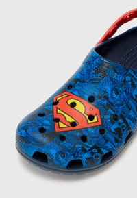 Blue rubber slip-on shoe featuring a prominent red and yellow Superman logo, with black comic-style graphic patterns and ventilation holes.