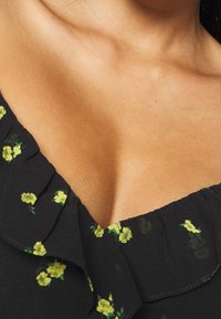 Nahaufnahme des Oberkörpers und Halses einer Frau, die ein schwarzes Kleid mit kleinen gelben Blumenmustern und einem rüschengeschnittenen Ausschnitt trägt.