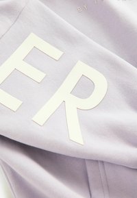 Tessuto lilla con grandi lettere bianche, dalla texture liscia e dal design informale. Le lettere sono leggermente in rilievo, aggiungendo dimensione.