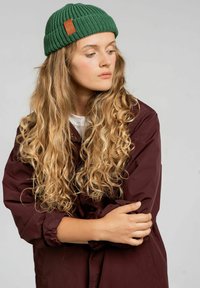 Zielona dzianinowa czapka z teksturowanym wzorem i brązową skórzaną metką. Modelka ma na sobie bordową kurtkę na białym topie, prezentując długie falowane włosy.