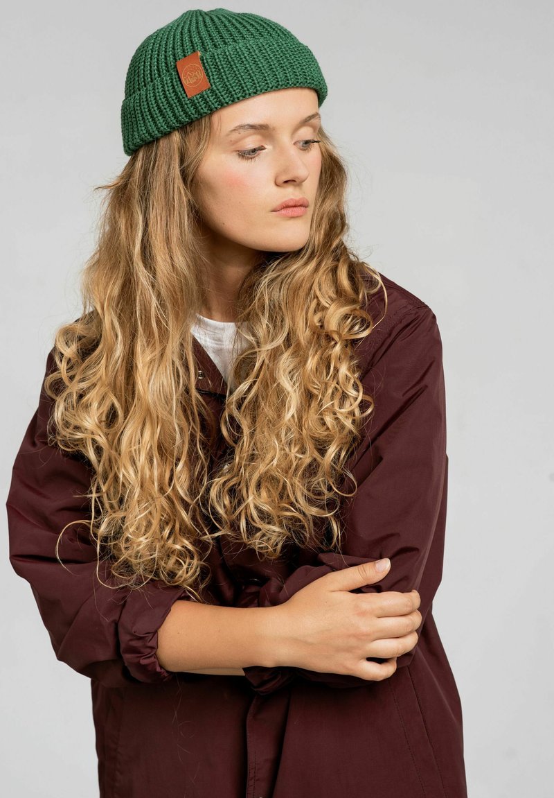 Zielona dzianinowa czapka z teksturowanym wzorem i brązową skórzaną metką. Modelka ma na sobie bordową kurtkę na białym topie, prezentując długie falowane włosy.