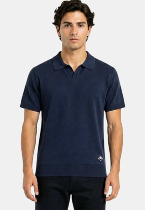 Giovane uomo che indossa una polo blu navy a maniche corte con un piccolo logo vicino all'orlo, in piedi davanti a uno sfondo semplice.