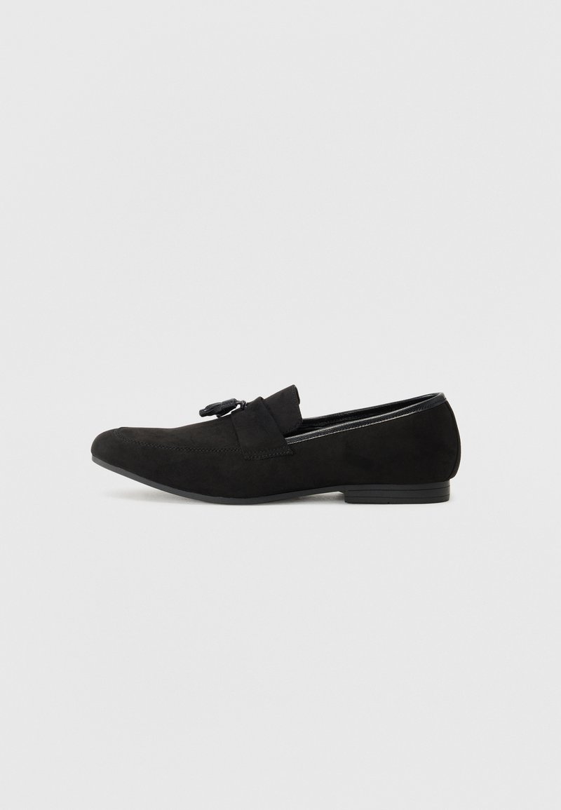 Mocassin en daim noir avec détail pompon, talon bas et design sans lacets, présenté de profil sur un fond clair uni.