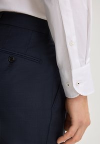 Witte blouse met lange mouwen en knoopmanchetten naast donkerblauwe textuurbroek met een gladde afwerking en een enkele knoopdetail.