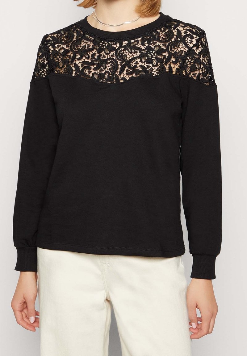 Felpa nera a maniche lunghe con sovrapposizione di pizzo sulle spalle, caratterizzata da un colletto rotondo e polsini a coste. Perfetta per look a strati.