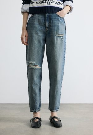 Personne portant un jean bleu délavé à taille haute avec des déchirures et des ourlets retroussés, ainsi que des chaussures Mary Jane noires, main dans la poche.