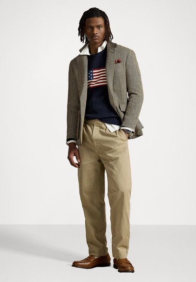 Blazer en chevrons marron, pull bleu marine avec un design de drapeau américain, chinos beiges et chaussures en cuir marron. Les accessoires notables incluent un pochette rouge.