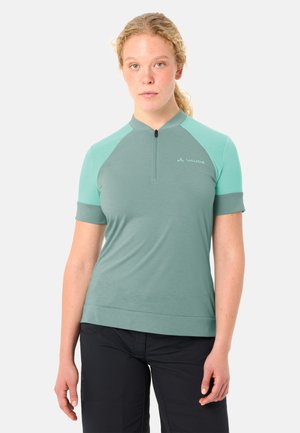 Giovane donna con capelli biondi ricci, indossa una maglietta sportiva a maniche corte verde chiaro con zip e pantaloni neri, in piedi contro uno sfondo bianco.